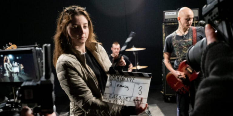 young filmmaker stands with a clapperboard in front of a camera. Behind her is a band ready to be filmed.