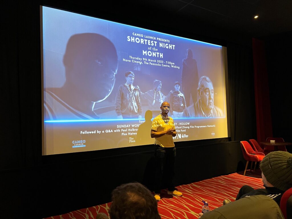 Person presents to an audience in front of a cinema screen displaying a holding slide.
