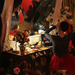A photo of a young woman sitting, looking in a large mirror putting red fabric in her hair. The dressing table is adorned with roses and bouquets.