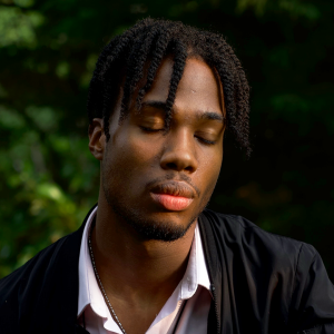 A young person with short braided hair and stubble. Their eyes are closed.